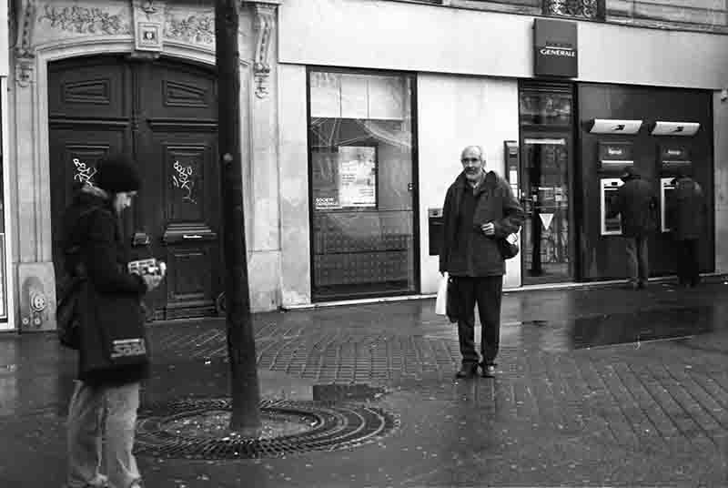 Black and white Paris street photography scene in front of a Société Générale bank branch on a wet city sidewalk, featuring an older man standing and looking towards a photographer holding a camera in the foreground.