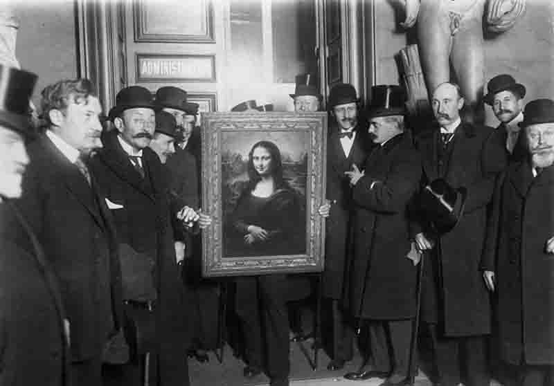 A black-and-white historical photograph shows a group of men in overcoats and hats, including top hats and bowlers, gathered around Leonardo da Vinci's 'Mona Lisa.' The painting is held upright in its ornate frame, positioned in the center of the group in front of a doorway labeled 'ADMINISTRATION.' To the right, a large classical statue is partially visible. The men, many with mustaches, are looking toward the painting or the camera in what appears to be a formal or official setting.